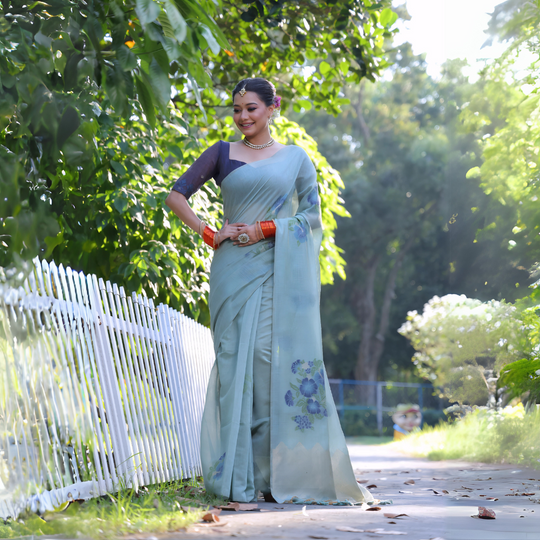 A woman models an elegant Sky Blue Soft Muga Cotton Saree. The pallu features large, artistic Resham floral motifs in shades of blue and indigo. She wears a contrasting navy blouse and is accessorized with a pearl choker necklace and vibrant orange bangles, posing outdoors by a white fence.