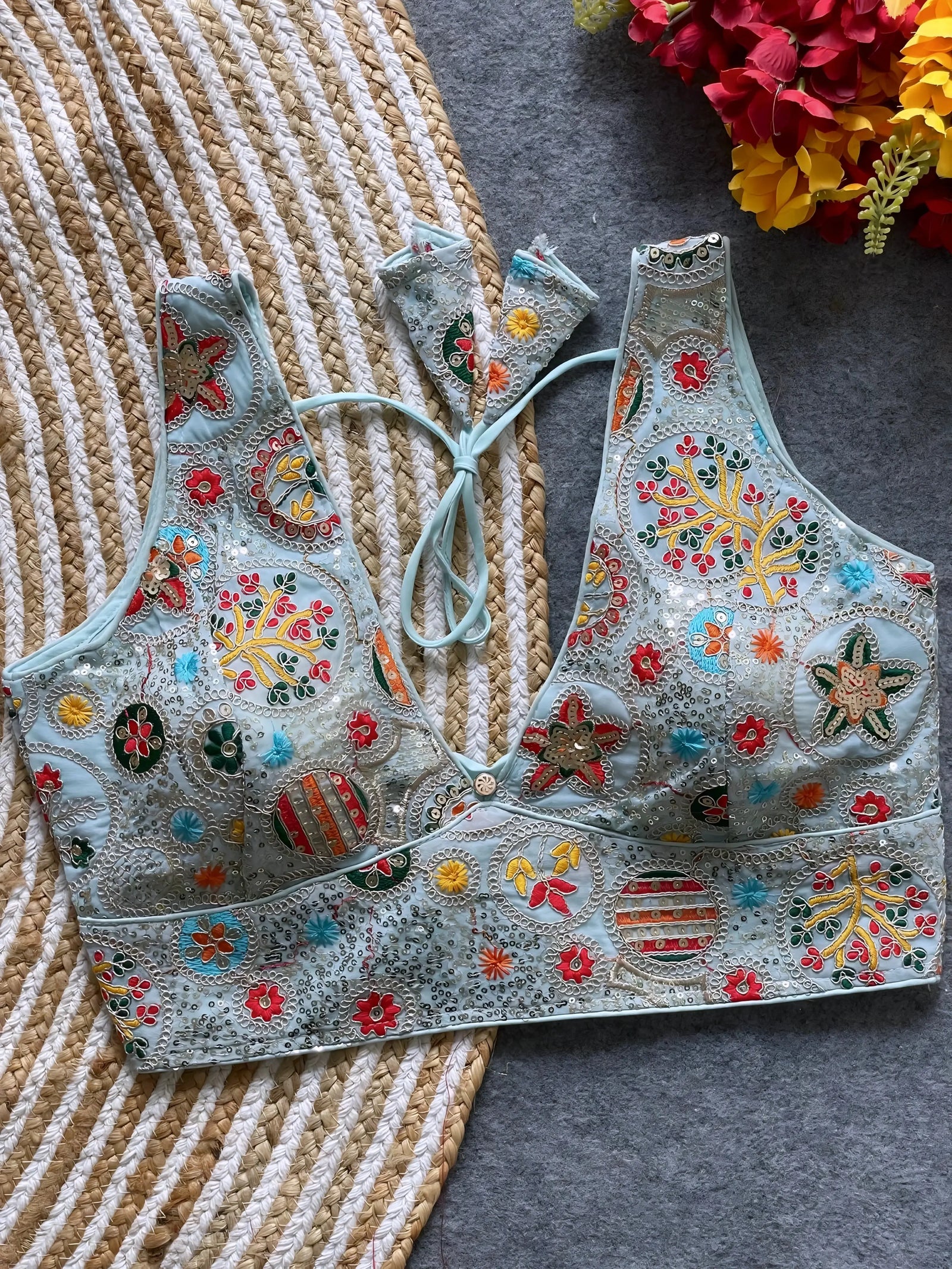  Pastel blue blouse featuring vibrant multicolor thread embroidery and mirror work in floral and geometric patterns, with sequin accents and a deep V-neck, displayed flat on a textured mat with artificial red and yellow flowers in the corner.