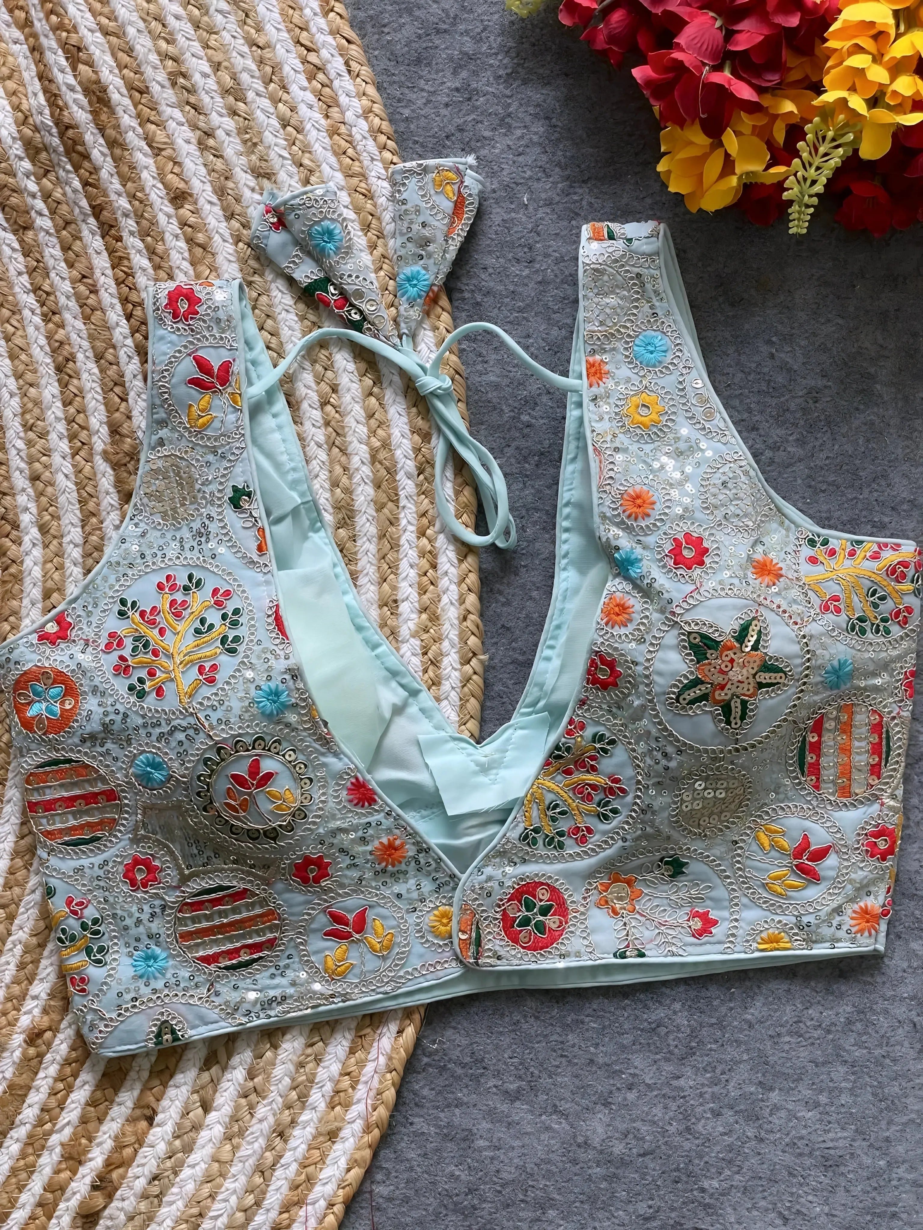  Pastel blue blouse featuring vibrant multicolor thread embroidery and mirror work in floral and geometric patterns, with sequin accents and a deep V-neck, displayed flat on a textured mat with artificial red and yellow flowers in the corner.
