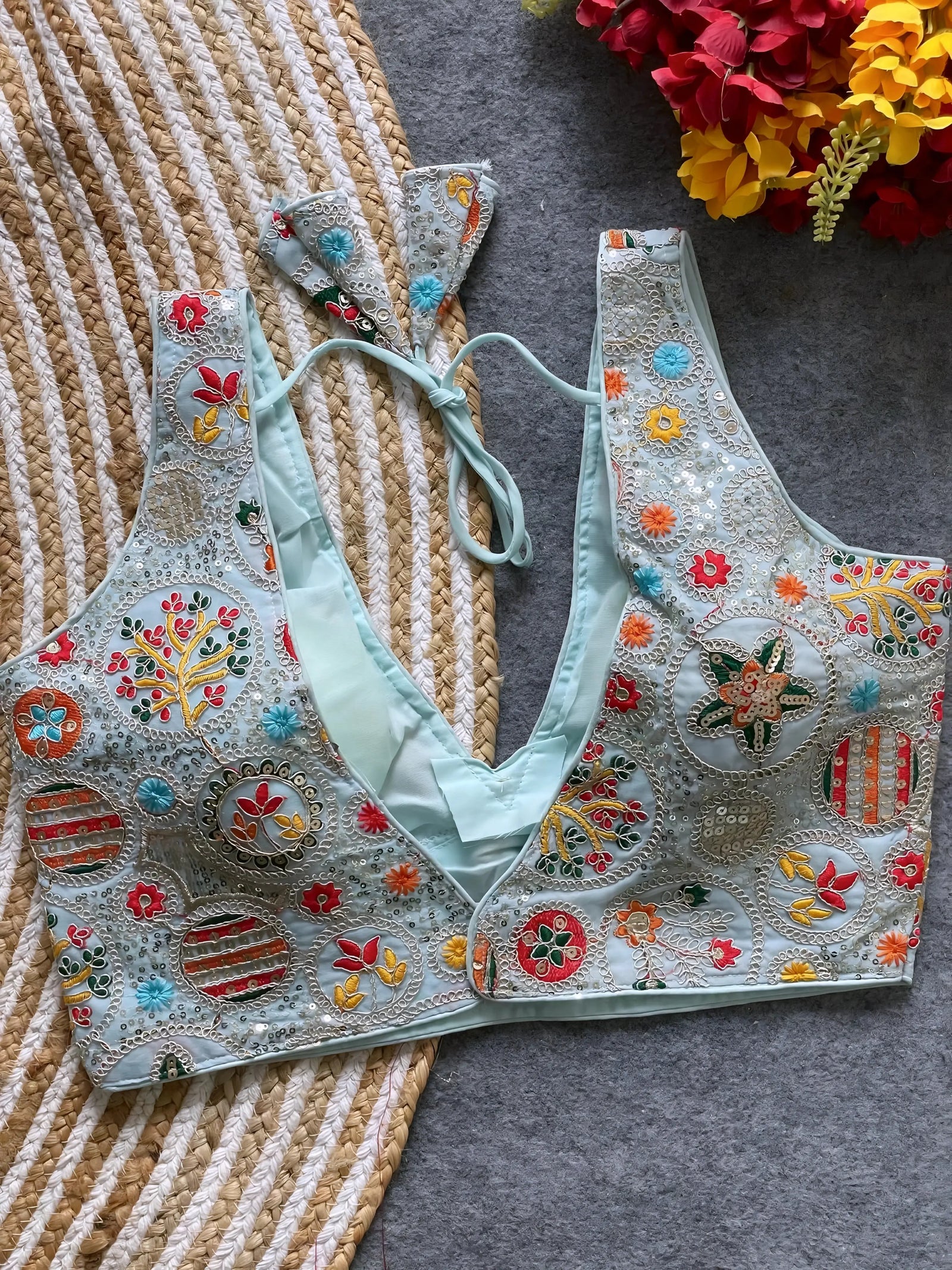  Pastel blue blouse featuring vibrant multicolor thread embroidery and mirror work in floral and geometric patterns, with sequin accents and a deep V-neck, displayed flat on a textured mat with artificial red and yellow flowers in the corner.