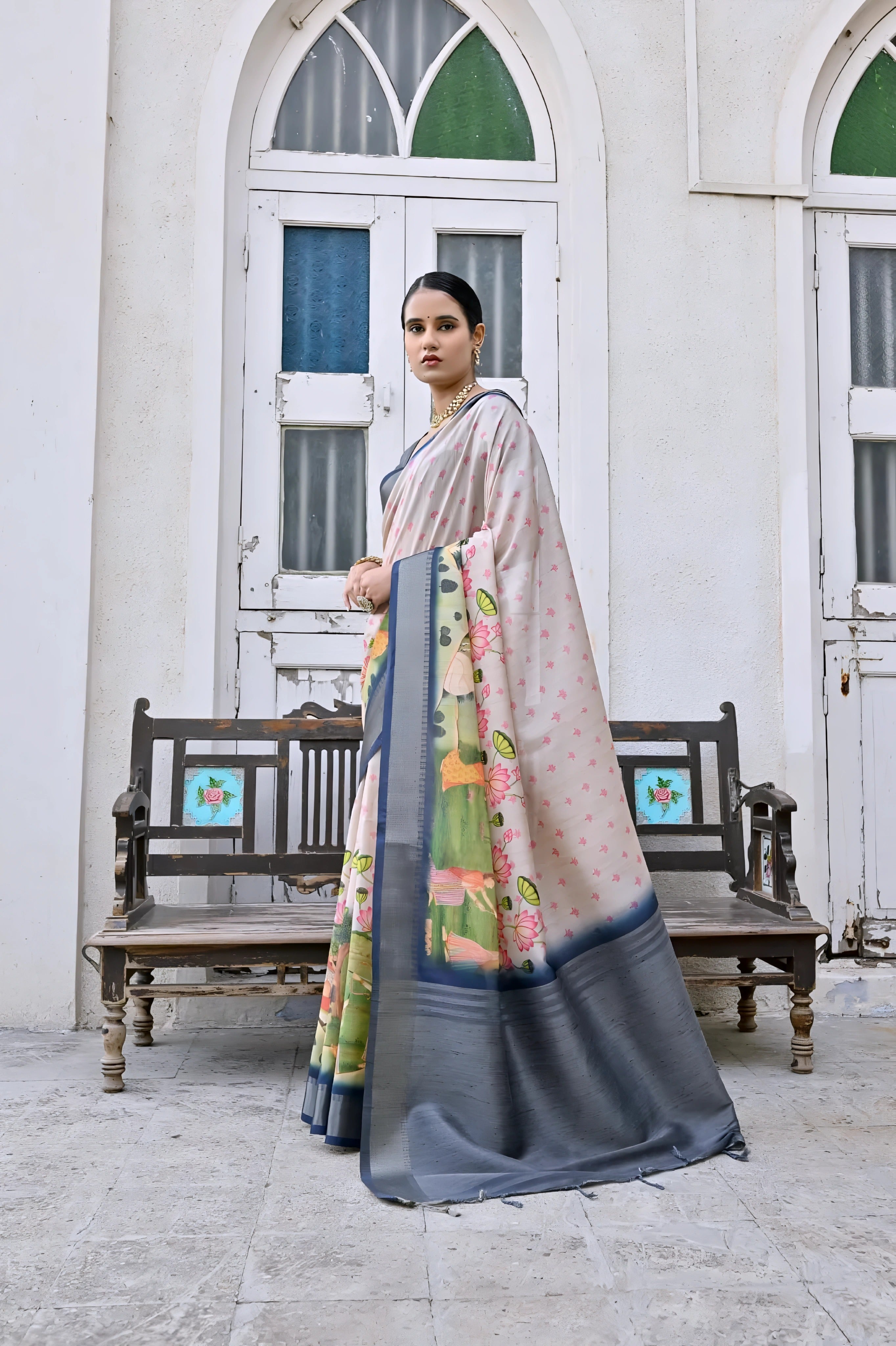 A woman models an elegant Off-White Soft Silk Saree featuring a large-scale floral and figurative Pichwai-style print in green and pink. The saree has a wide Slate Grey/Navy Blue border and pallu. She wears a matching dark grey blouse and a gold Kundan choker necklace, posing outdoors next to an old wooden bench.