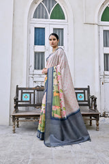 A woman models an elegant Off-White Soft Silk Saree featuring a large-scale floral and figurative Pichwai-style print in green and pink. The saree has a wide Slate Grey/Navy Blue border and pallu. She wears a matching dark grey blouse and a gold Kundan choker necklace, posing outdoors next to an old wooden bench.