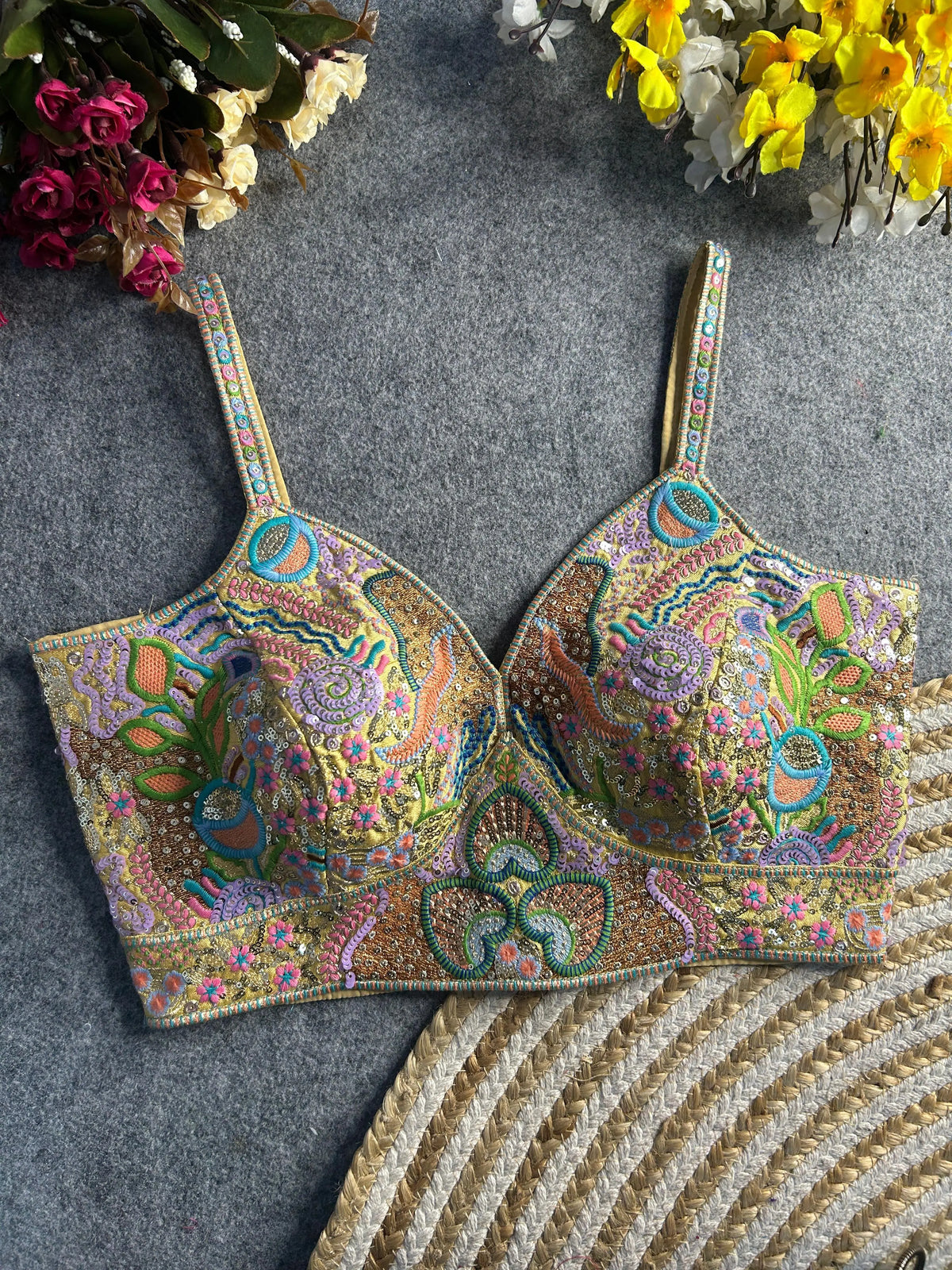  Pastel multicolor bustier-style blouse featuring intricate thread embroidery and sequin embellishments in floral, paisley, and geometric motifs, with structured cups and adjustable straps, displayed flat on a textured grey surface surrounded by artificial pink, yellow, and white flowers in the corners.