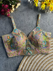  Pastel multicolor bustier-style blouse featuring intricate thread embroidery and sequin embellishments in floral, paisley, and geometric motifs, with structured cups and adjustable straps, displayed flat on a textured grey surface surrounded by artificial pink, yellow, and white flowers in the corners.