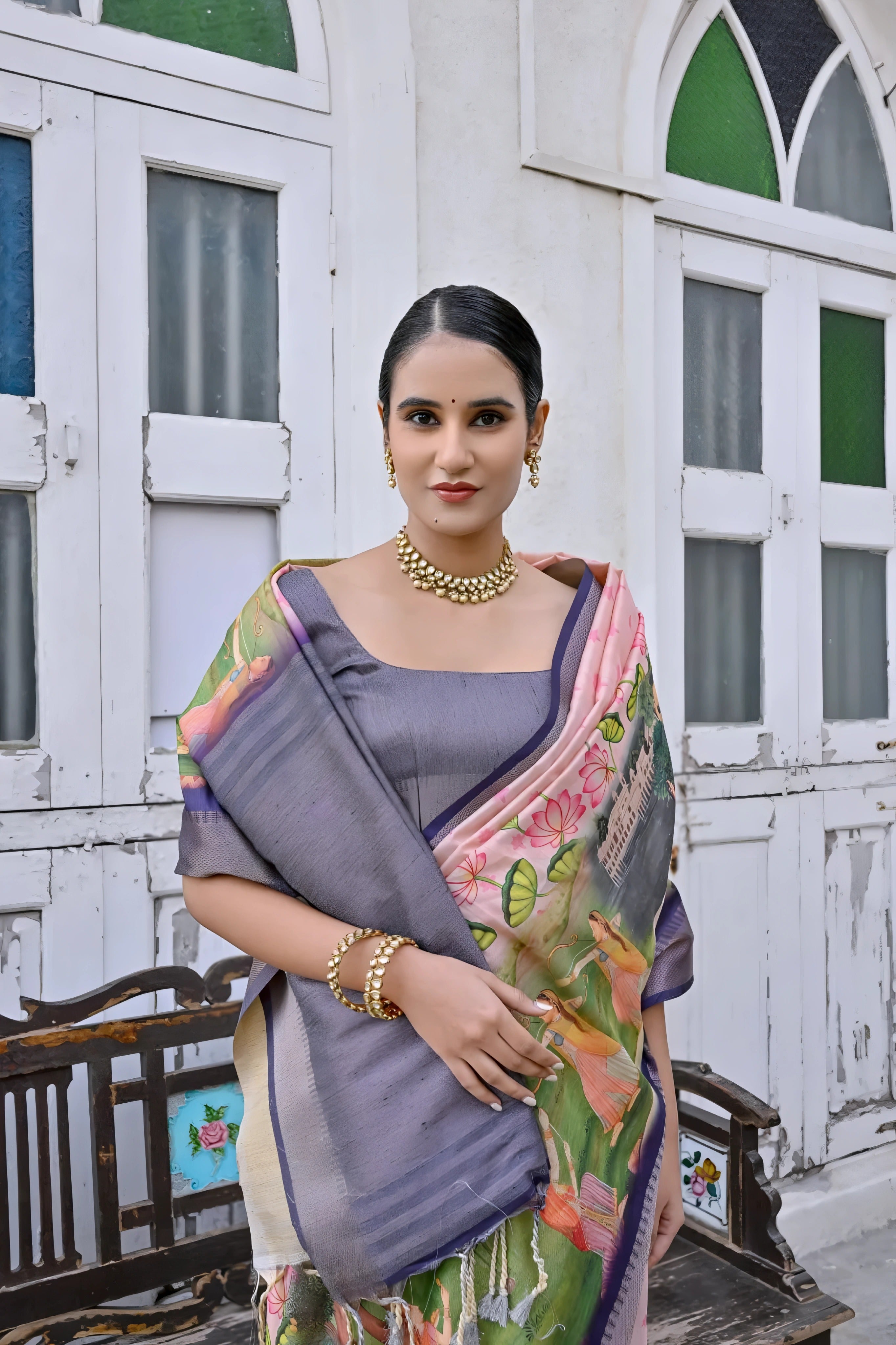 A woman models an elegant Light Peach Soft Silk Saree featuring a large-scale floral and figurative Pichwai-style print. The saree has a wide Slate Grey/Smoky Purple border and pallu. She wears a matching short-sleeve grey blouse and a gold Kundan choker necklace, posing outdoors next to an old wooden bench.