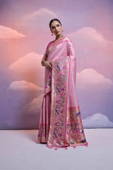 A woman models a graceful Soft Pink Banarasi Silk Saree. The saree features a wide, vibrant Deep Pink border and pallu with dense, colorful floral and vine Kanjivaram-style weaving. She wears a matching short-sleeve pink blouse and a gold choker necklace, posing indoors against a dreamy cloud backdrop.