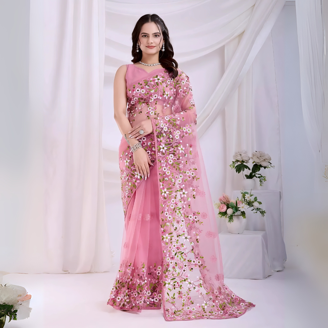 A woman models an ethereal Light Pink Soft Net Saree. The sheer fabric is densely covered in delicate all-over floral embroidery featuring white, pink, and green threadwork. She wears a matching sleeveless pink V-neck blouse and accessorizes with sophisticated silver/diamond jewelry, including a layered necklace and cuff bracelet.