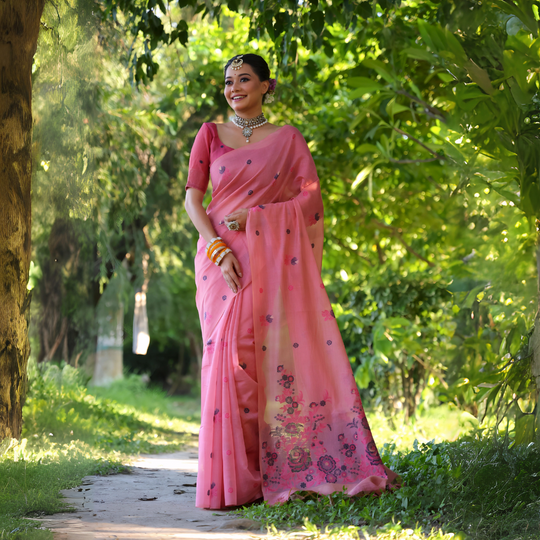 A woman models an elegant light Pink Soft Muga Cotton Saree. The body has scattered floral Resham weaving, and the pallu features an elaborate panel of darker floral weaving/print in purple and blue tones. She wears a matching pink blouse, white stone/Kundan jewelry, and vibrant orange and yellow bangles, posing outdoors.