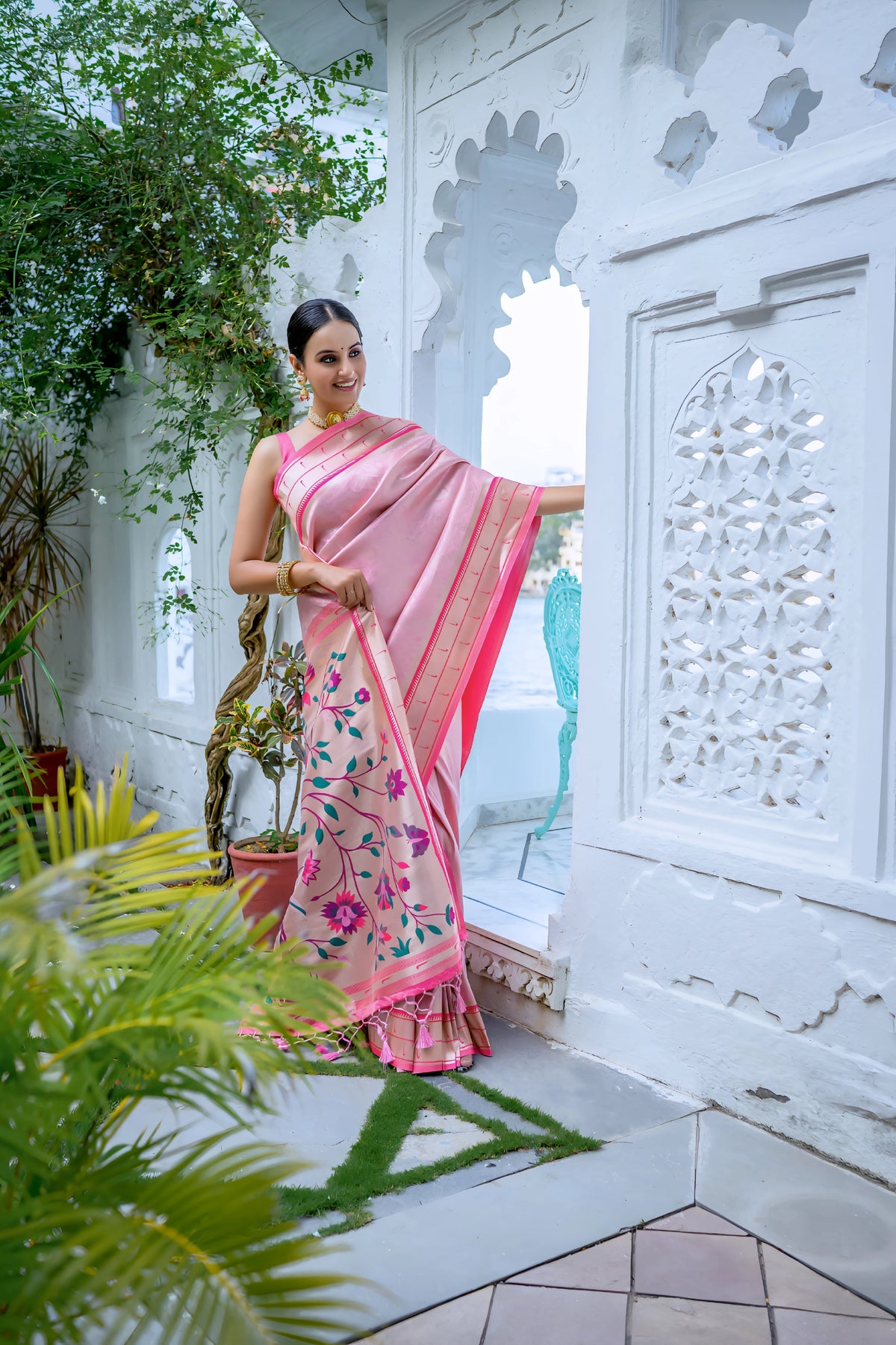 A woman models an elegant Soft Pink Silk Saree. The saree features a golden Zari border with a dark pink contrast piping. The pallu is a focal point, featuring rich, artistic Paithani-style print or weaving of pink, red, and brown bird-on-branch motifs. She wears a pink sleeveless blouse and traditional gold jewelry, posing in a white, ornate courtyard.