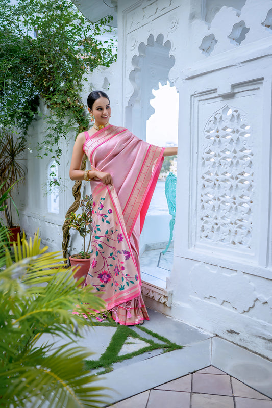 A woman models an elegant Soft Pink Silk Saree. The saree features a golden Zari border with a dark pink contrast piping. The pallu is a focal point, featuring rich, artistic Paithani-style print or weaving of pink, red, and brown bird-on-branch motifs. She wears a pink sleeveless blouse and traditional gold jewelry, posing in a white, ornate courtyard.