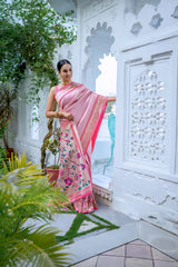 A woman models an elegant Soft Pink Silk Saree. The saree features a golden Zari border with a dark pink contrast piping. The pallu is a focal point, featuring rich, artistic Paithani-style print or weaving of pink, red, and brown bird-on-branch motifs. She wears a pink sleeveless blouse and traditional gold jewelry, posing in a white, ornate courtyard.
