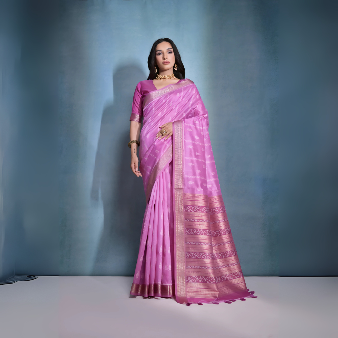 A woman models a stunning Deep Pink Soft Raw Silk Saree. The saree features subtle tone-on-tone Ikat weaving on the body, framed by a wide, luxurious border of dark magenta and intricate gold Zari weaving. She wears a matching pink blouse and gold Kundan jewelry, posing indoors against a textured blue backdrop.