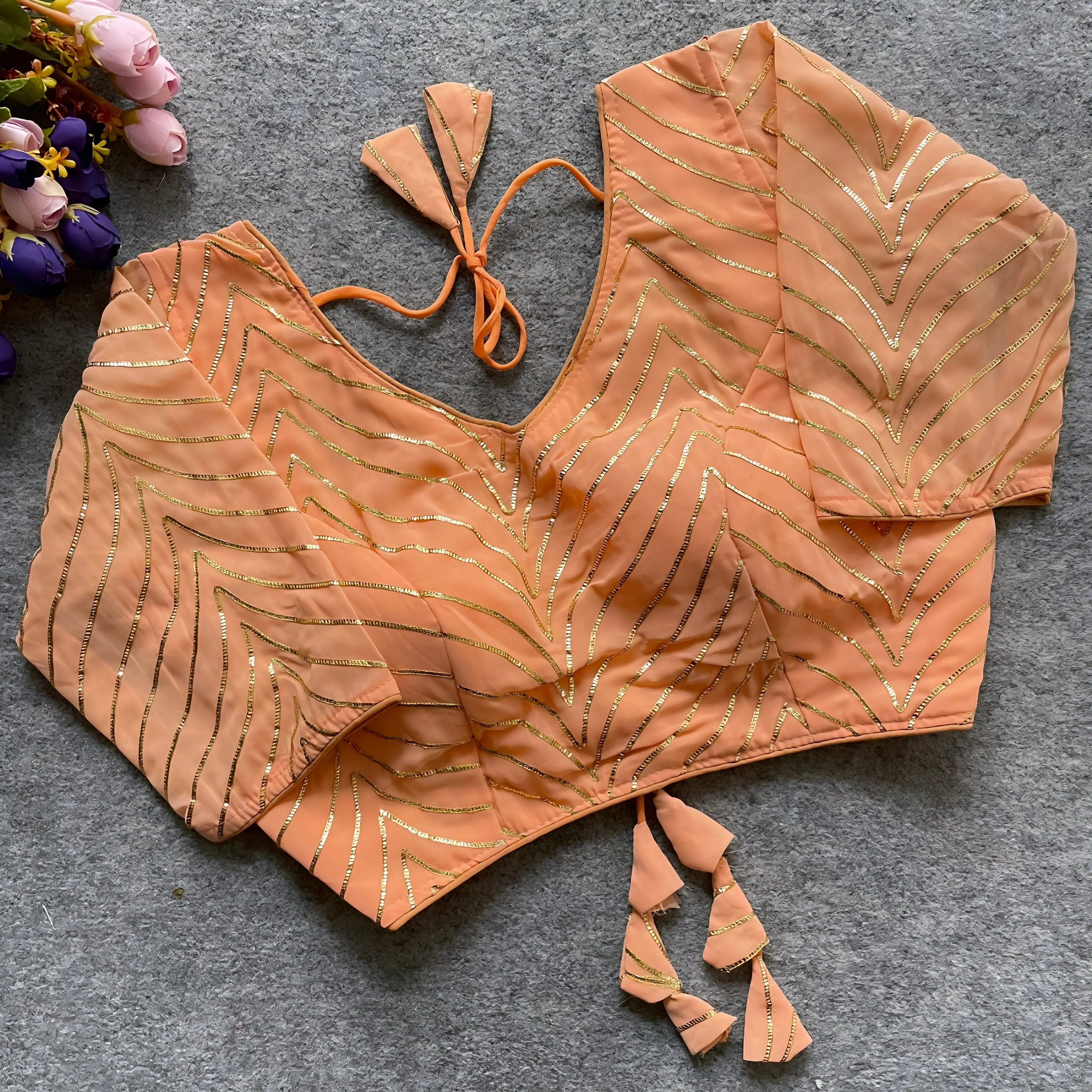 Peach blouse featuring elegant gold gota patti zigzag embroidery, three-quarter sleeves, and a stylish back tie-up with coordinated triangular tassels. Displayed flat on a grey textured surface beside pink and purple flowers, highlighting its festive and chic ethnic appearance.