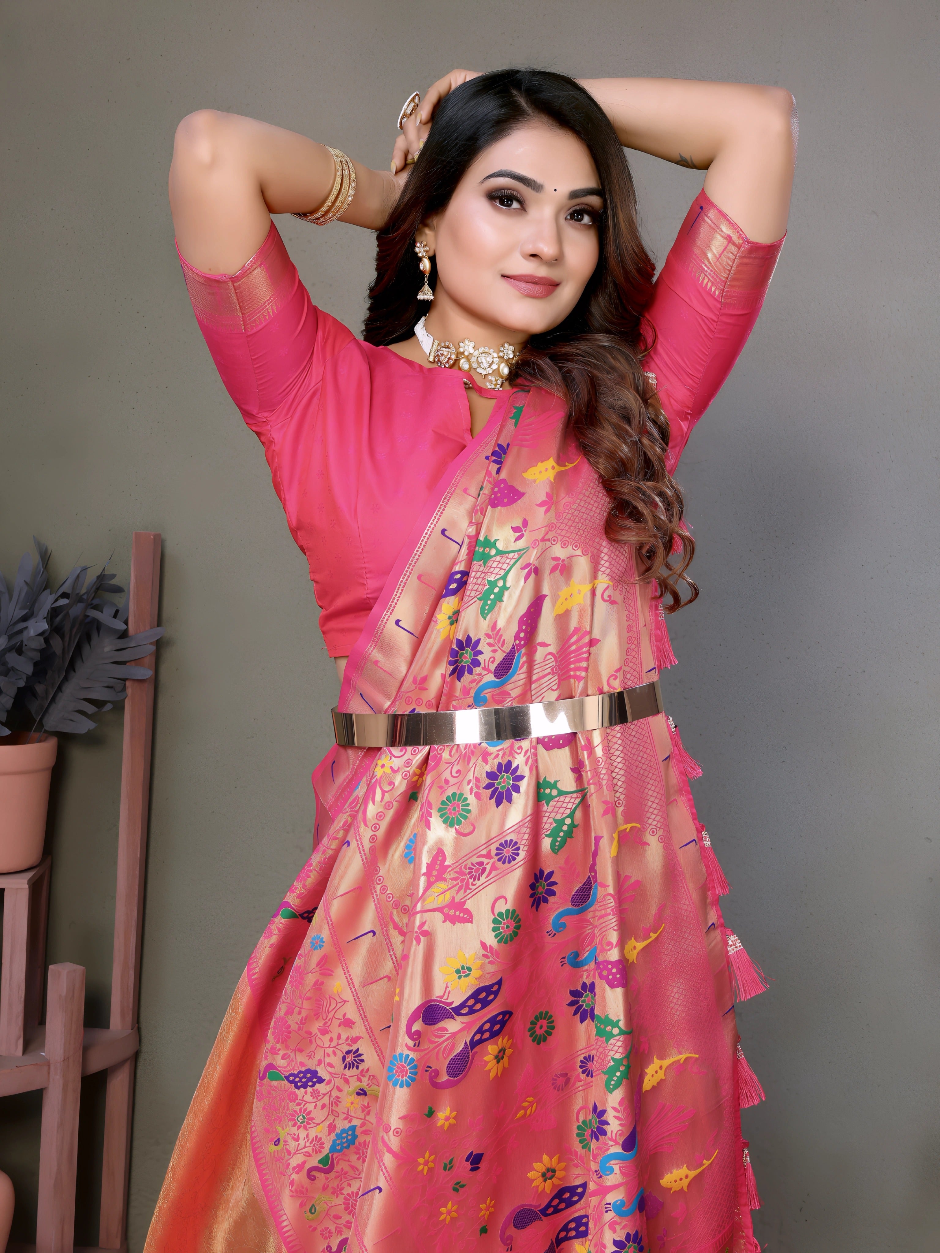 Woman wearing an elegant peach-pink silk saree with a bright pink blouse and intricate multicolor floral motifs on the pallu, finished with a shiny pink border. She is accessorized with a statement choker necklace, bangles, and rings, standing gracefully in a modern studio decorated with minimal wooden stands holding terracotta pots and green potted plants against a muted backdrop.
