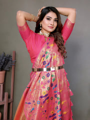 Woman wearing an elegant peach-pink silk saree with a bright pink blouse and intricate multicolor floral motifs on the pallu, finished with a shiny pink border. She is accessorized with a statement choker necklace, bangles, and rings, standing gracefully in a modern studio decorated with minimal wooden stands holding terracotta pots and green potted plants against a muted backdrop.