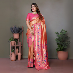 Woman wearing an elegant peach-pink silk saree with a bright pink blouse and intricate multicolor floral motifs on the pallu, finished with a shiny pink border. She is accessorized with a statement choker necklace, bangles, and rings, standing gracefully in a modern studio decorated with minimal wooden stands holding terracotta pots and green potted plants against a muted backdrop.