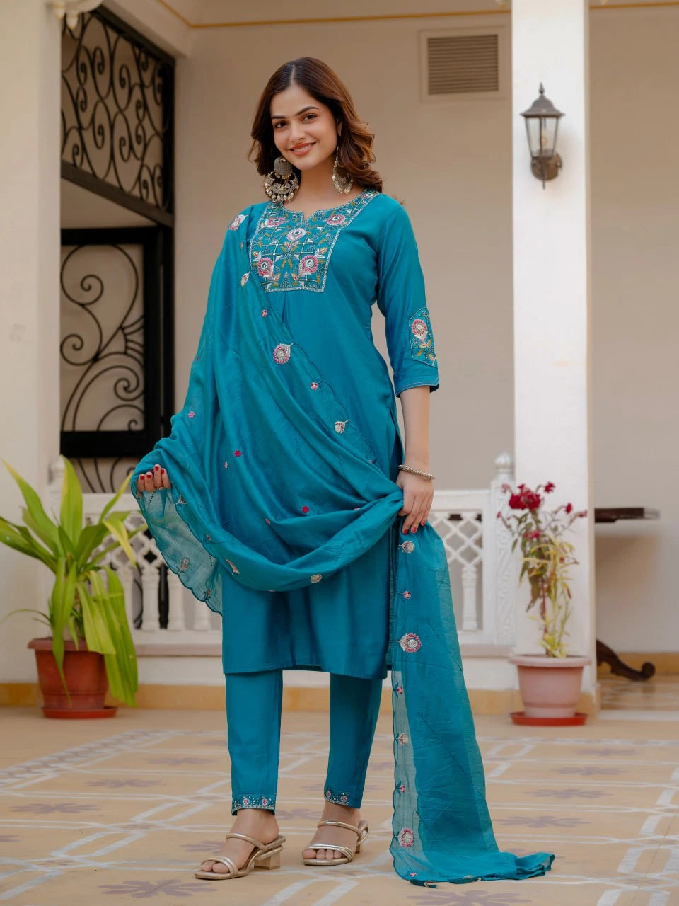 Woman wearing a blue embroidered outfit standing in a well-lit room with plants and furniture.