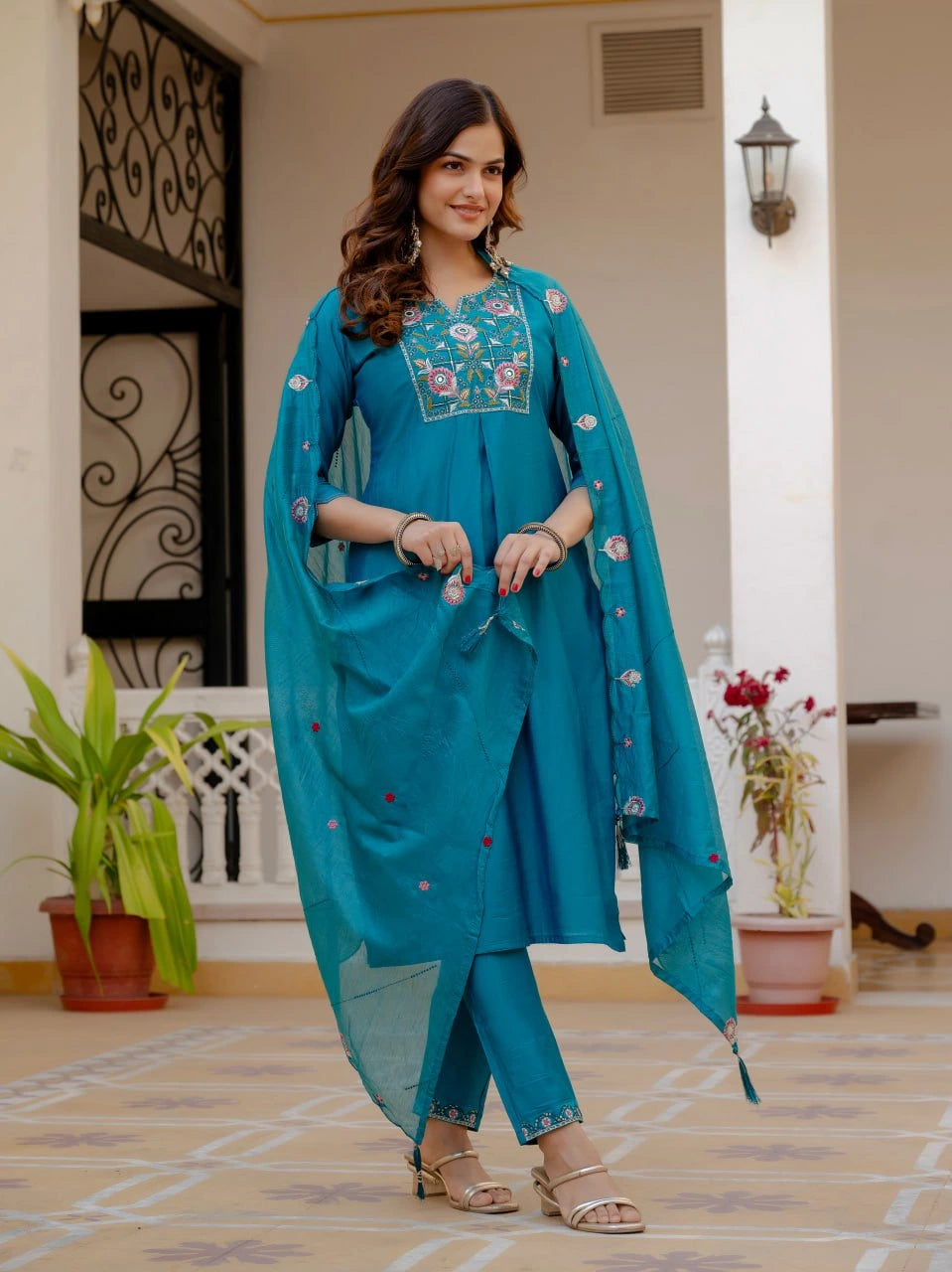 Woman in a teal traditional outfit with floral embroidery standing in a well-lit room.