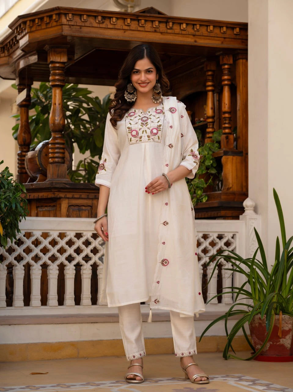 Woman in a white embroidered outfit standing in a decorative indoor setting with plants.