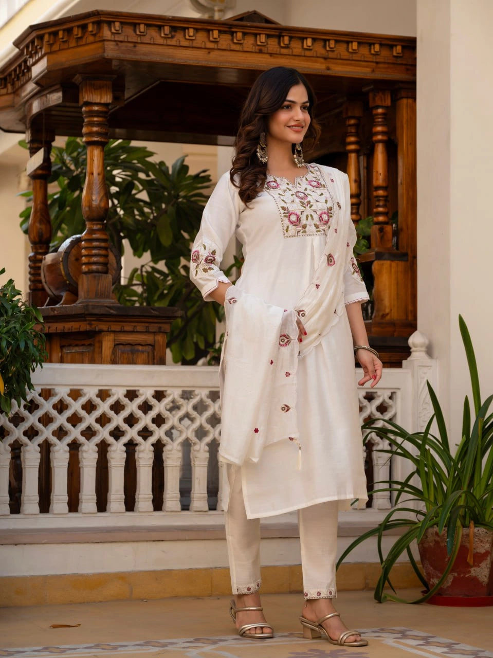 Woman in a white embroidered outfit standing in an elegant indoor setting with wooden decor and plants.