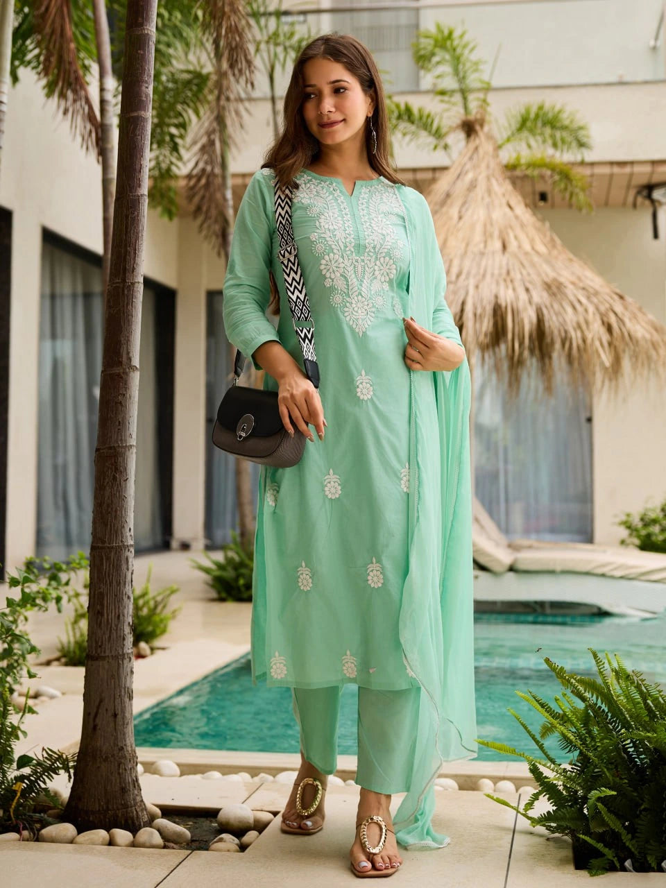 Woman in a light green traditional outfit standing by a poolside with palm trees in the background