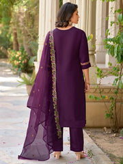 Woman in a purple traditional outfit standing outdoors with greenery around.