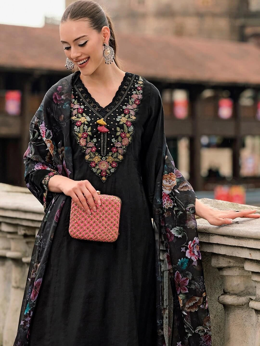 Woman wearing a black dress with floral embroidery and a floral shawl, holding a pink clutch.
