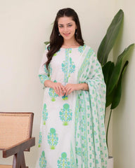 Woman wearing a white dress with green floral patterns, standing indoors with a plant in the background.