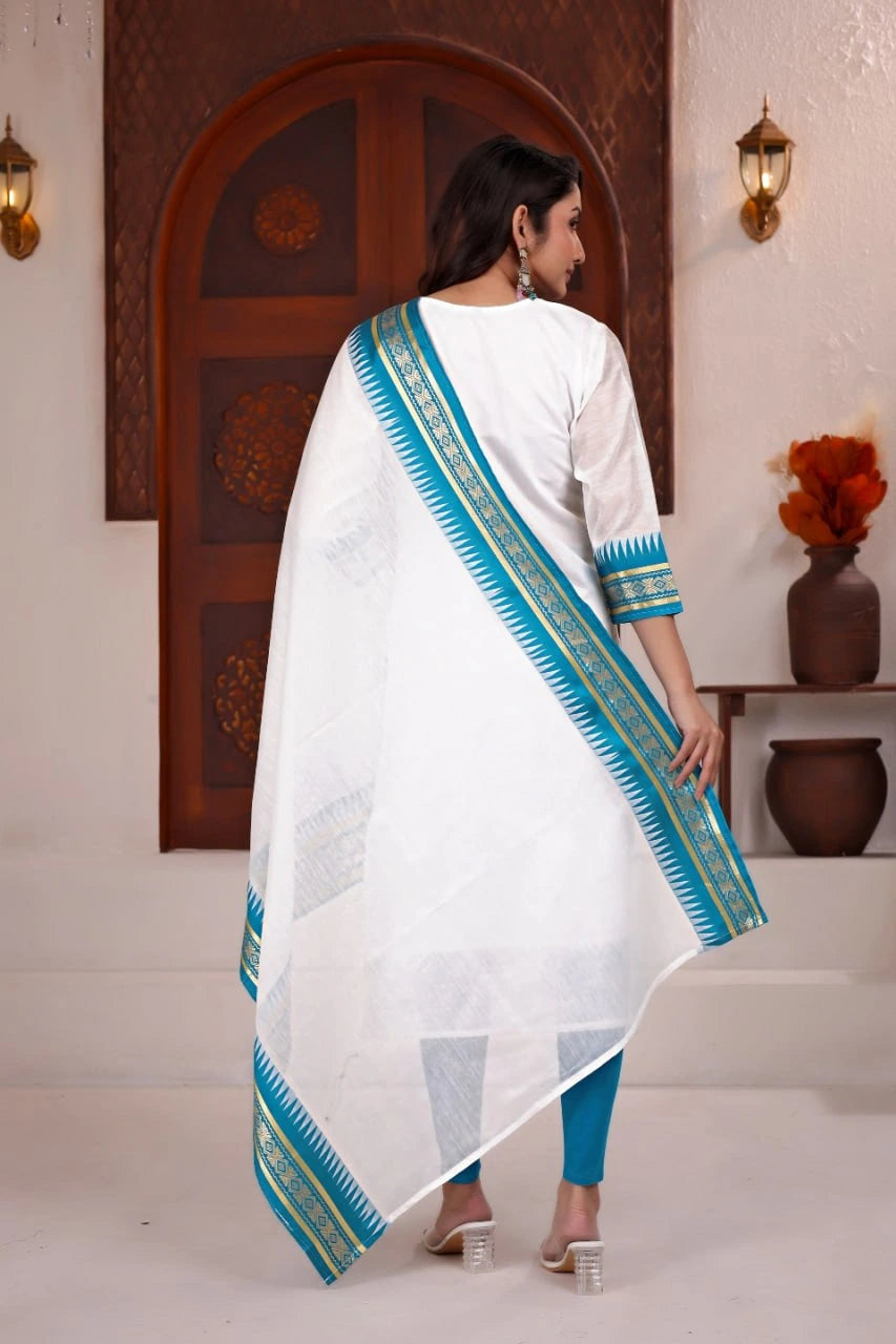 Woman wearing a white and blue traditional outfit with a decorative border in a room setting.