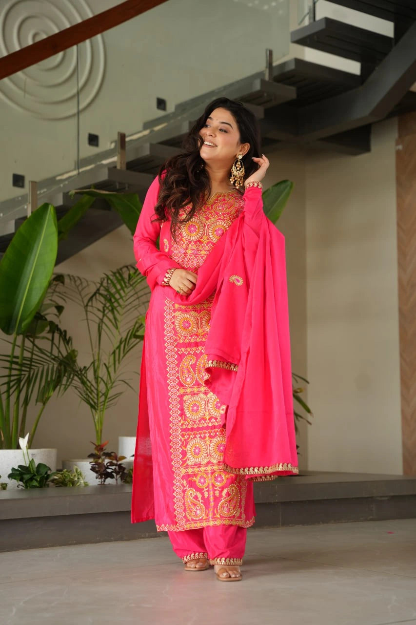 Woman in a bright pink traditional outfit with gold embroidery standing indoors.