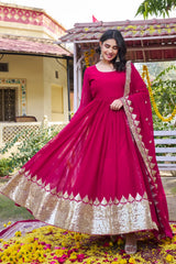Woman in a pink traditional outfit with gold details standing in front of a house with greenery.