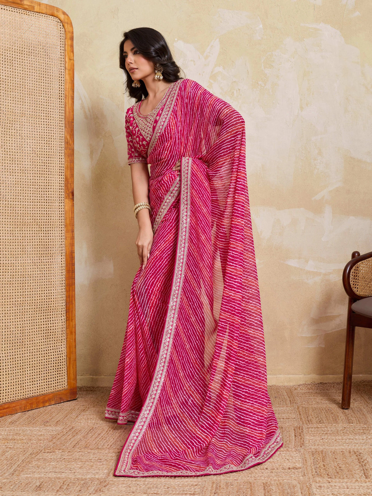 Woman wearing a pink saree with a white border in a room with a textured wall and wooden chair.