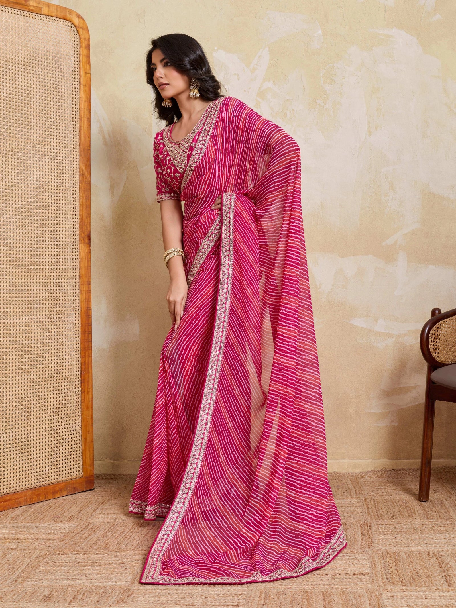 Woman wearing a pink saree with a white border in a room with a textured wall and wooden chair.