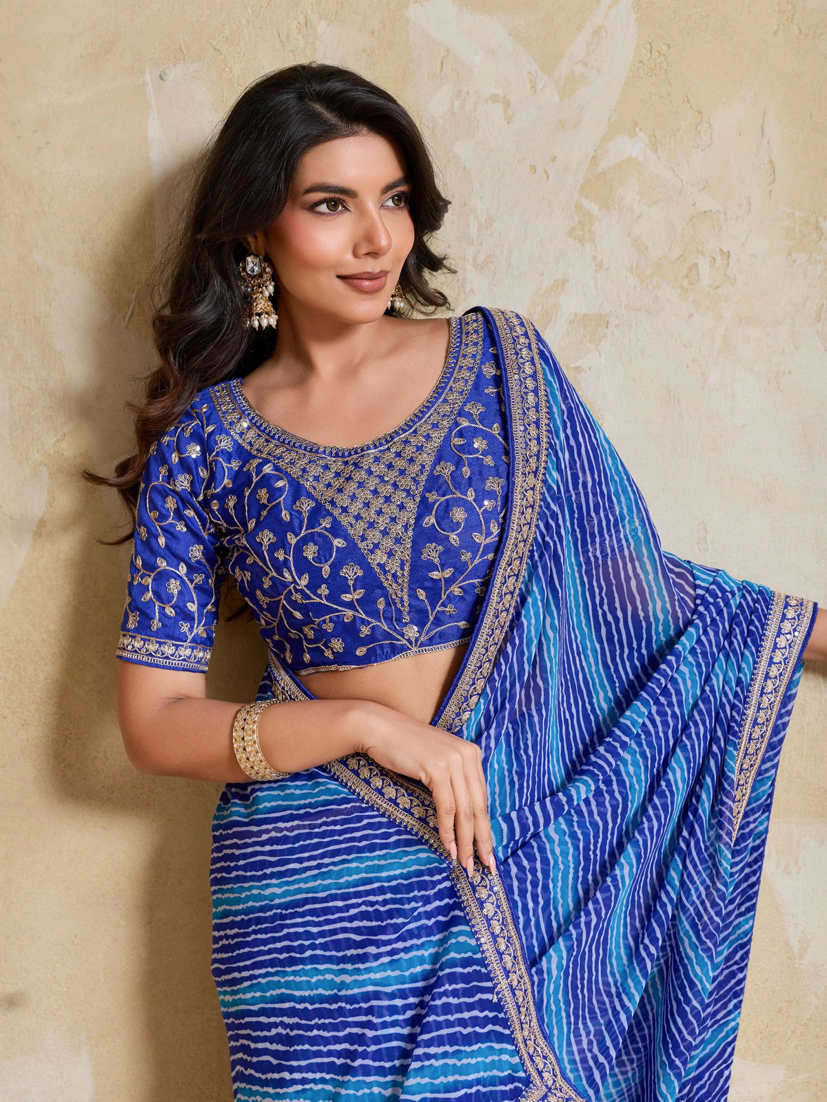 Woman wearing a blue saree with intricate patterns against a beige wall.
