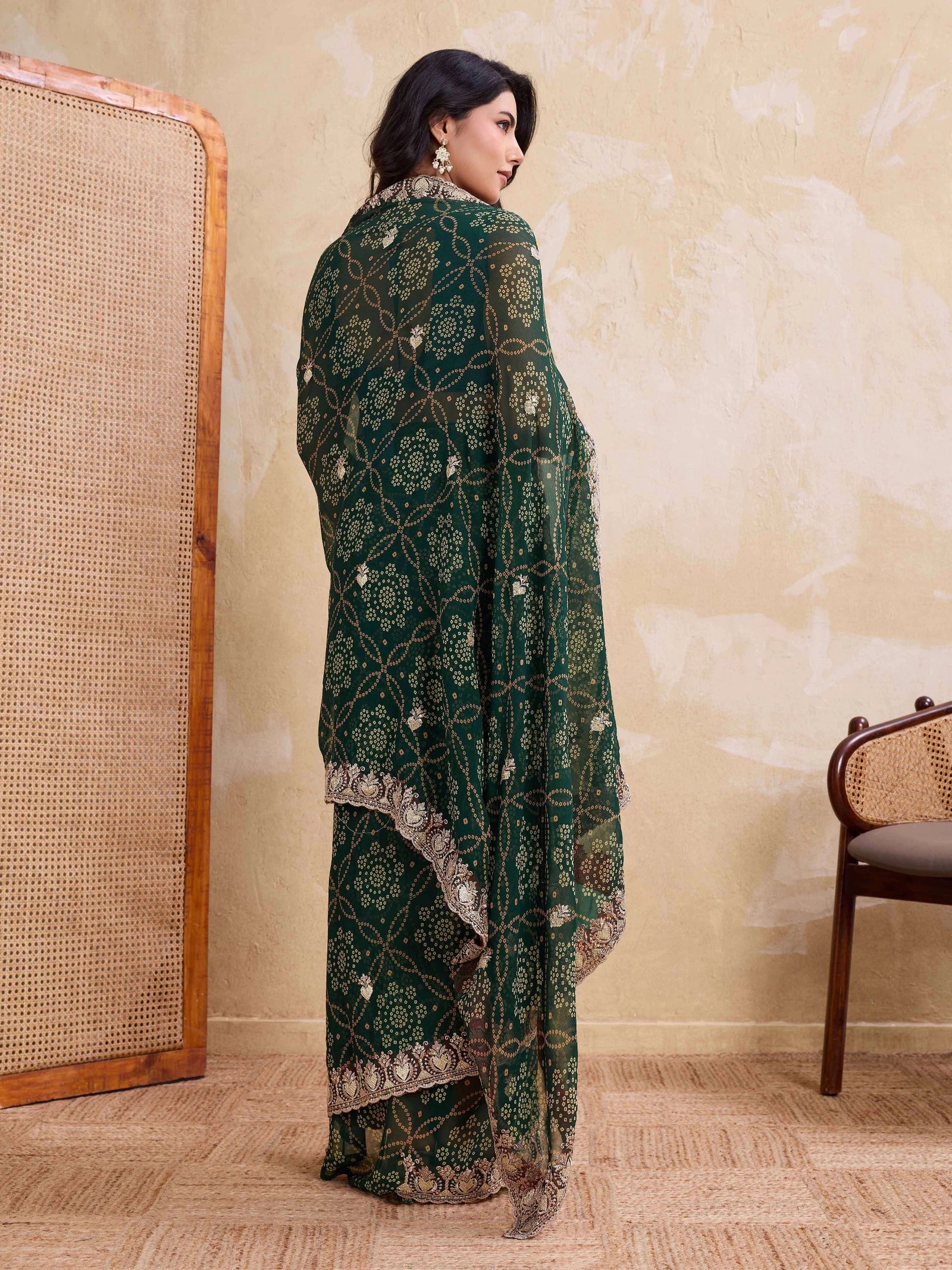 Woman in a green embroidered traditional outfit standing in a room with a textured wall and wooden chair.