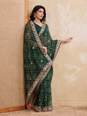 Woman wearing a green saree with silver border against a textured beige wall.