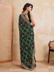 Woman wearing a green saree with a pattern, standing in a room with a textured wall and wooden chair.