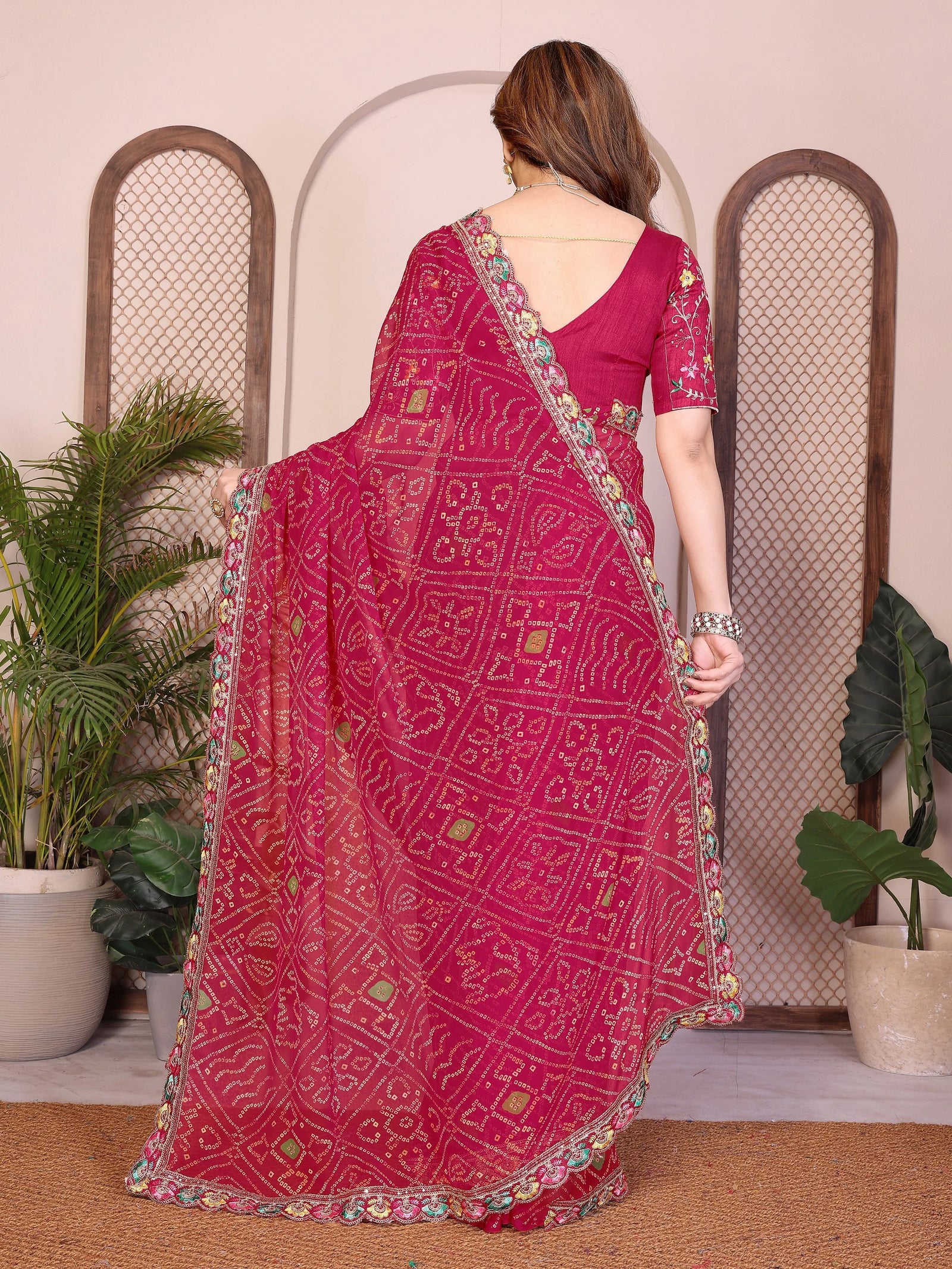 Woman wearing a pink saree with intricate patterns in a room with plants and a decorative arch.