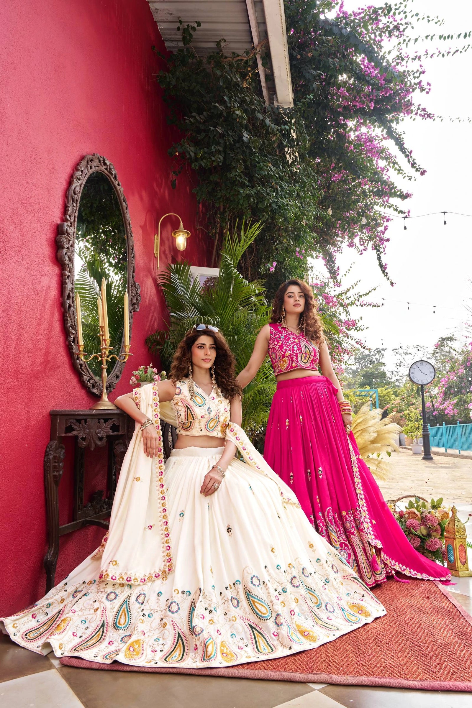 Two women in traditional outfits standing against a red wall with greenery.