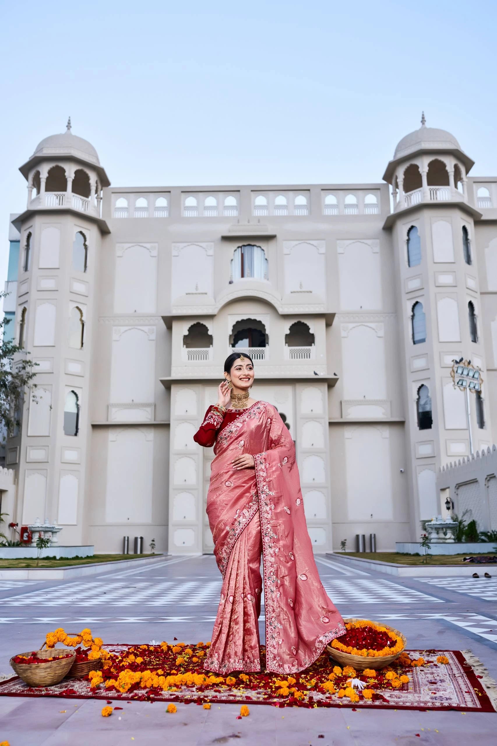 Woman in a pink saree standing in front of a building with decorative elements on the ground.