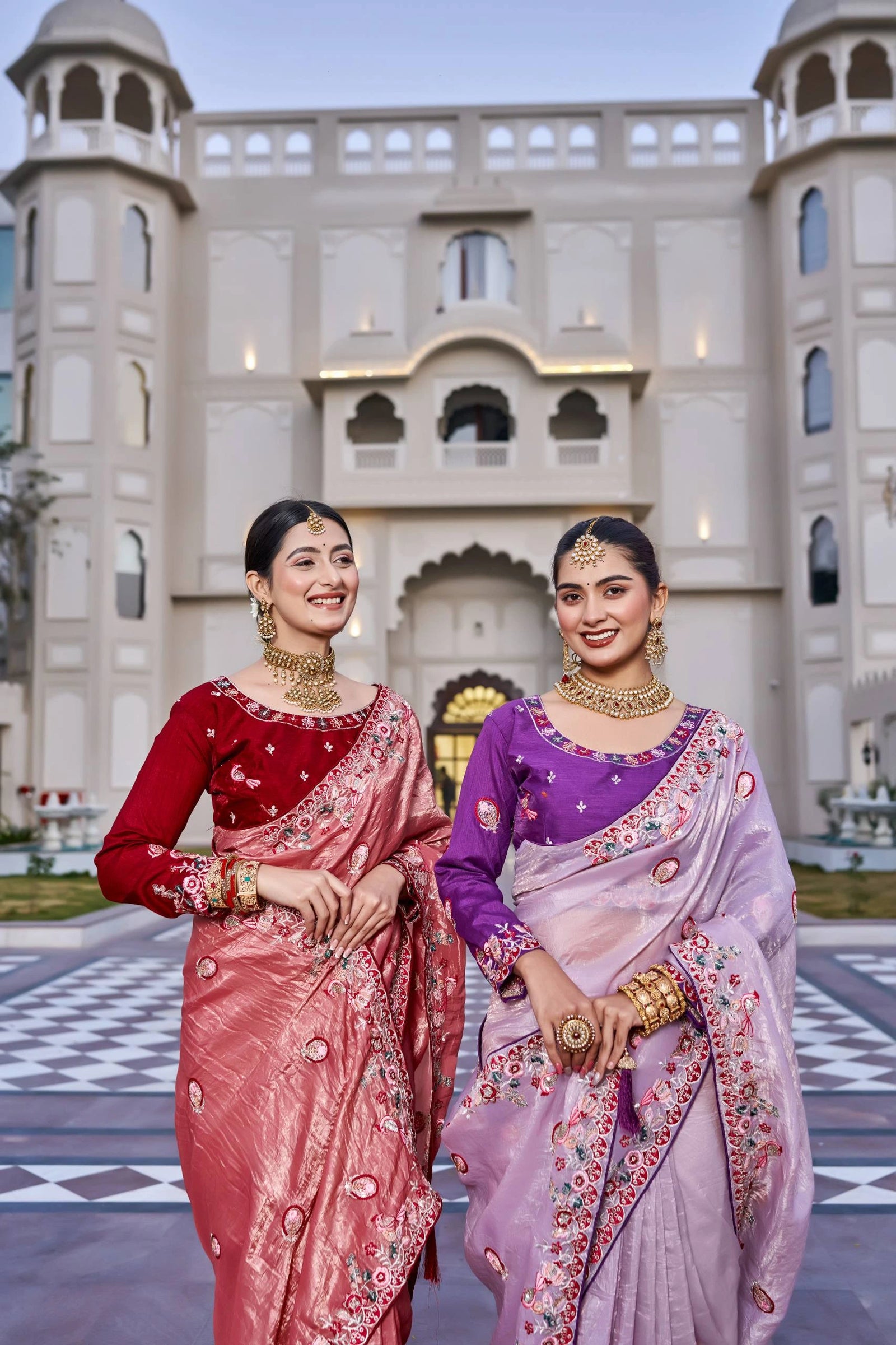 Two women in traditional sarees standing in front of an architectural building.