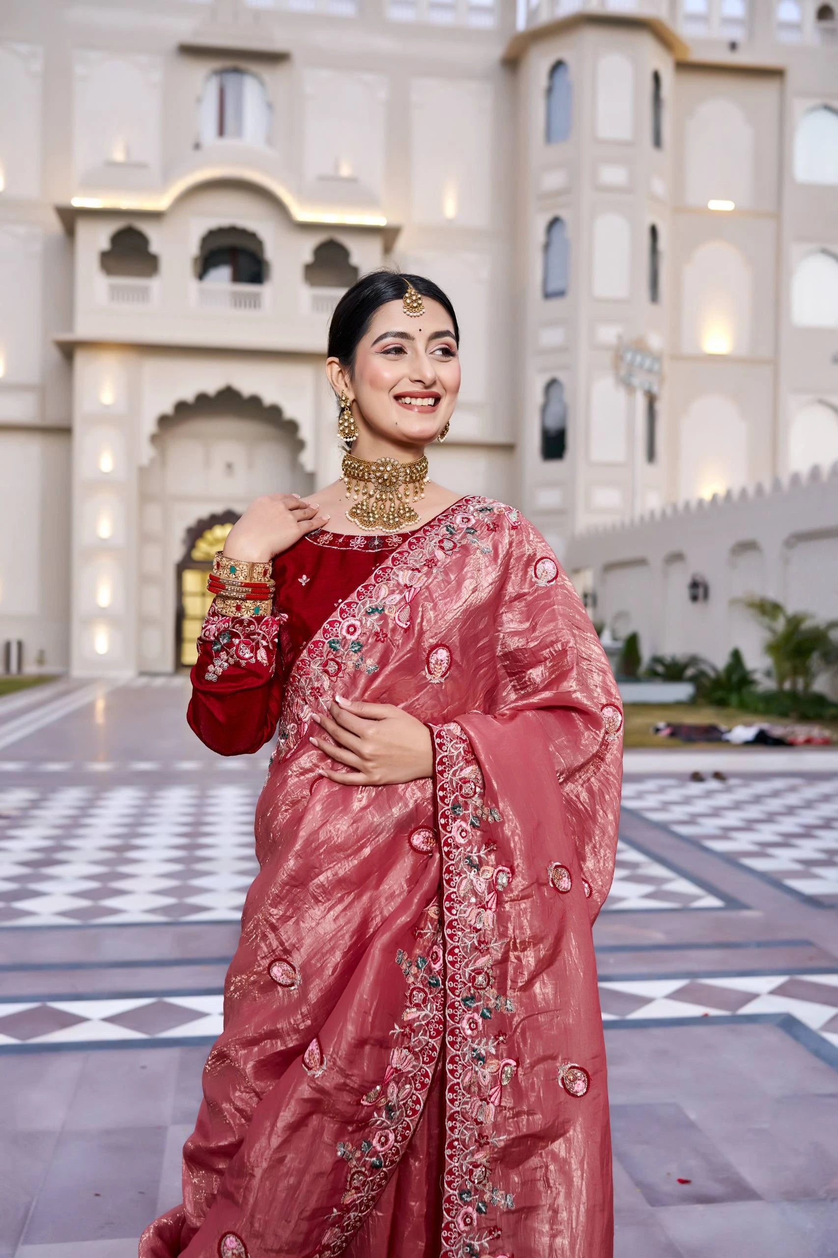 Woman in a pink saree with floral patterns standing in front of an architectural building.