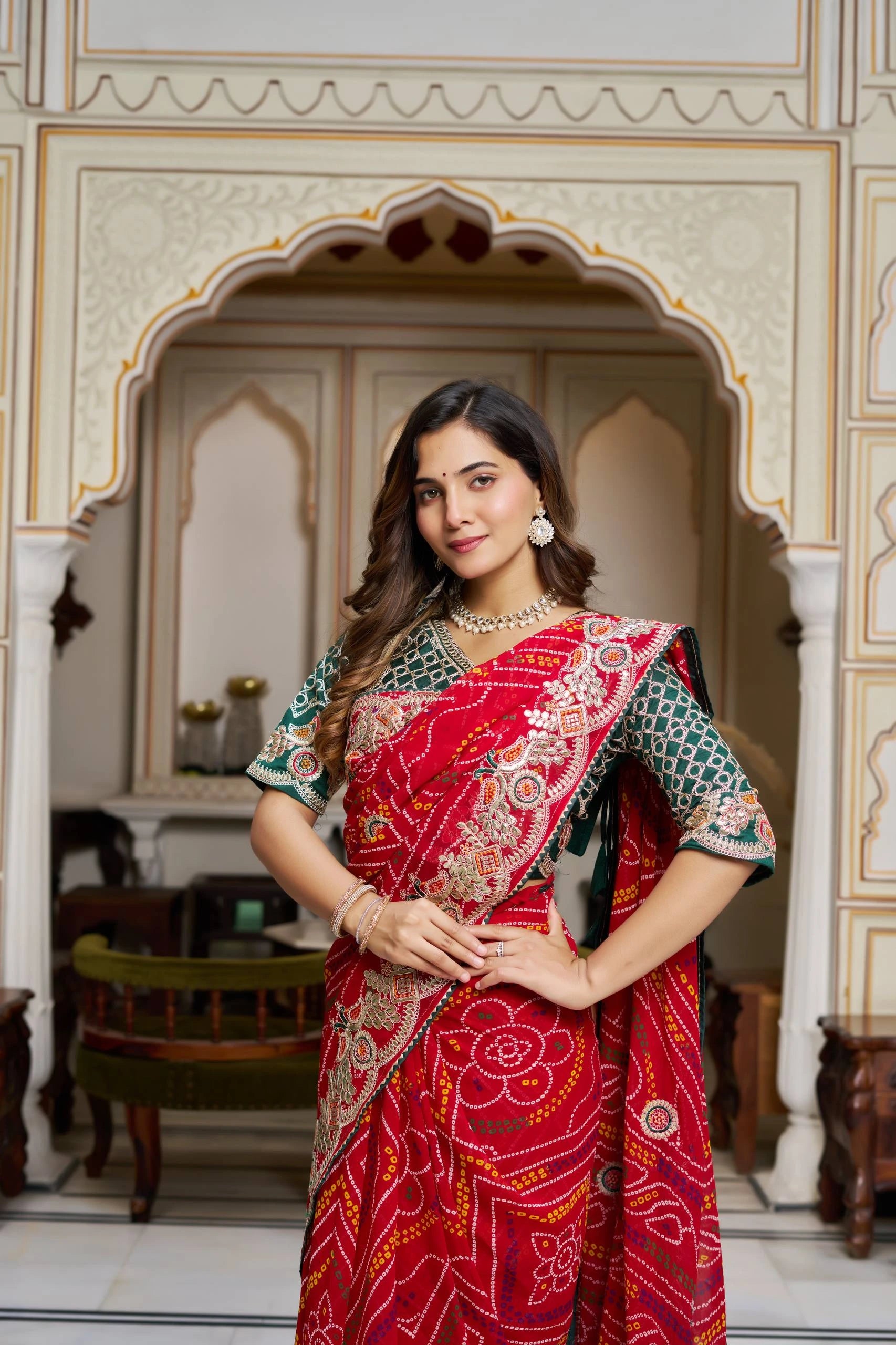 Woman in a red saree with intricate patterns standing in an ornate architectural setting.