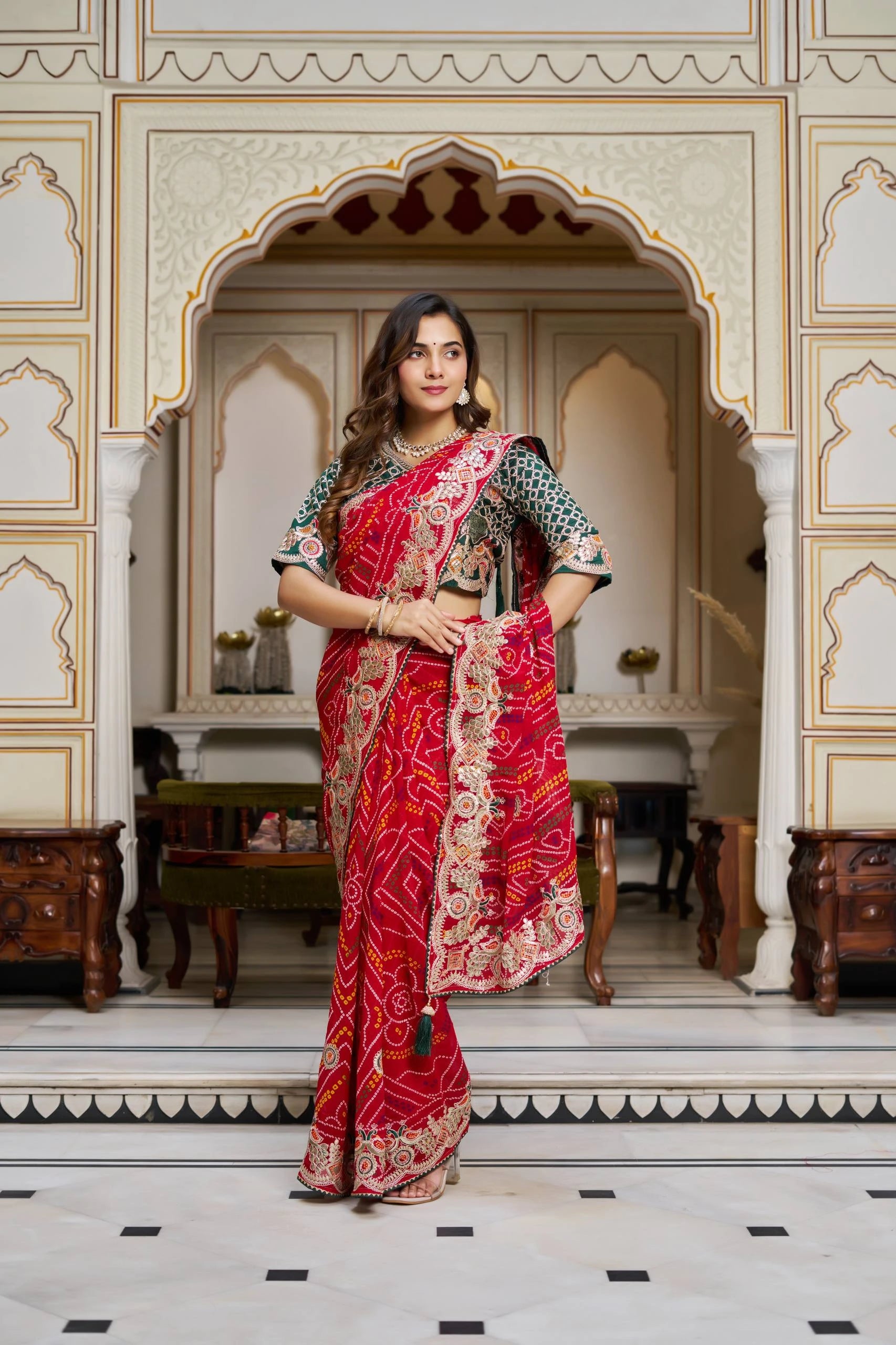 Woman in a red saree with intricate patterns standing in an ornate room.
