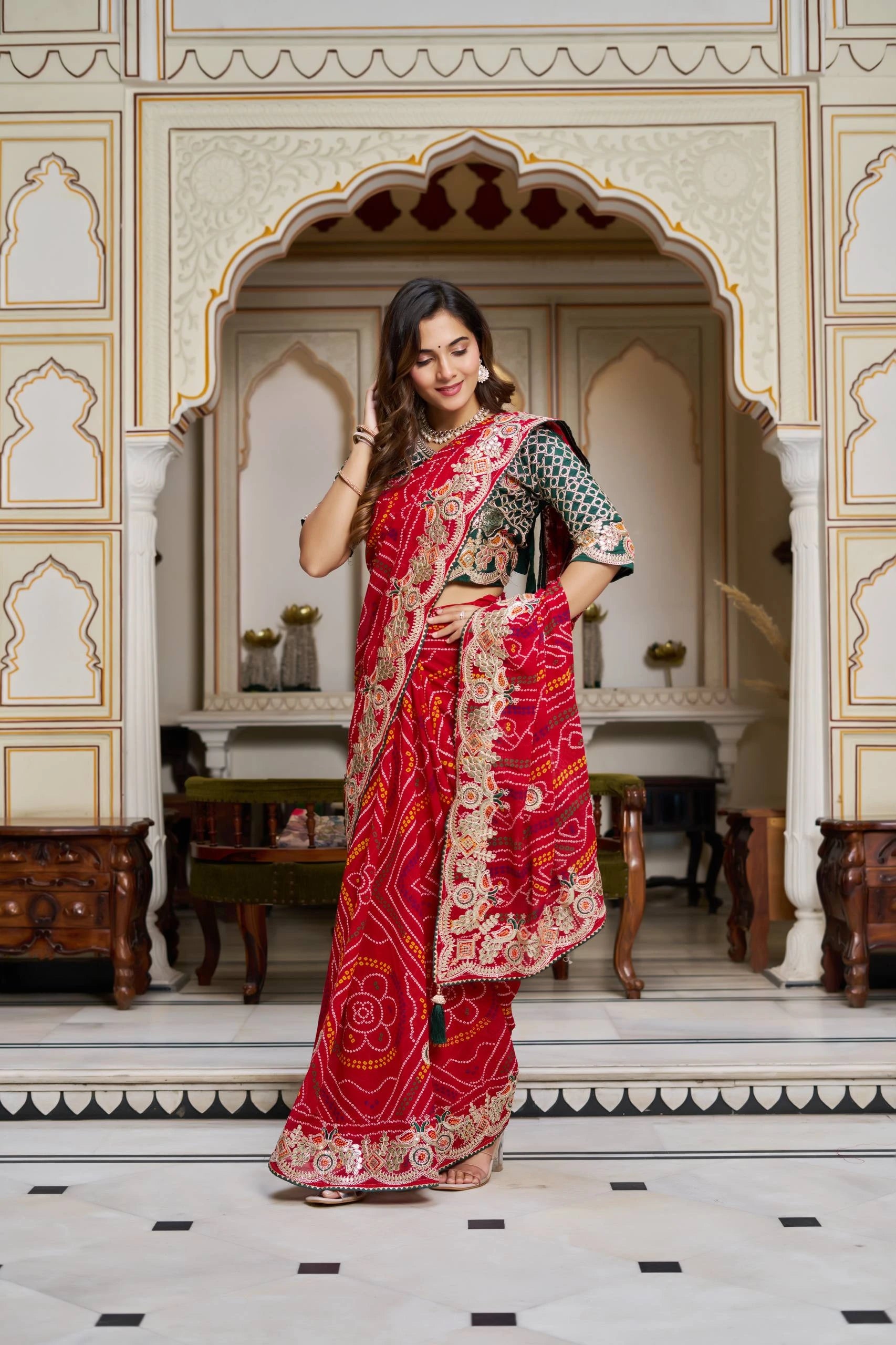 Woman in a red saree with gold patterns standing in an ornate room.