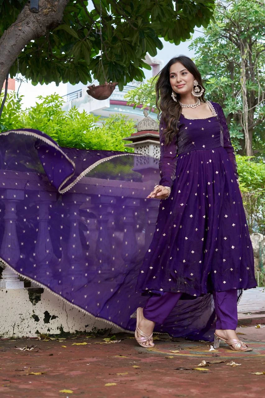 Woman in a purple traditional outfit with a long train, standing outdoors with trees and buildings in the background.
