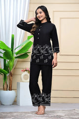 Woman wearing a black outfit with white patterns standing in a room with a plant and decorative items.