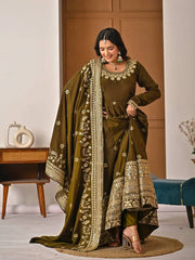 Woman in traditional embroidered outfit standing indoors with a wooden door and table in the background.