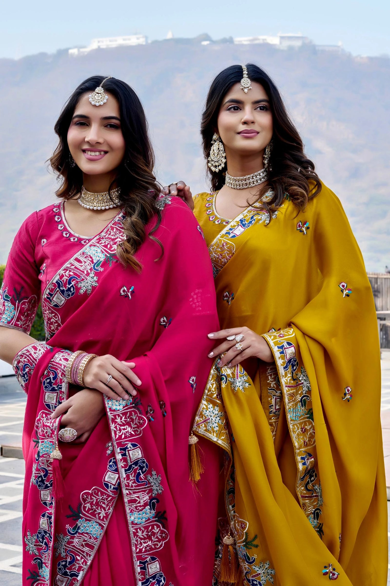 Two women in traditional pink and yellow sarees with mountainous background