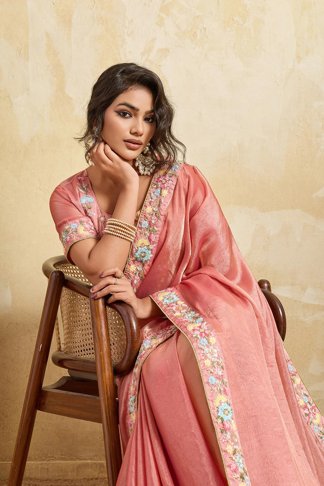 Woman in a pink saree with floral patterns sitting on a wooden chair against a beige wall.